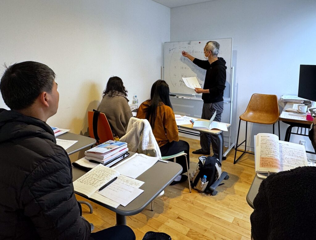 教室での講義。講師がホワイトボードに かな を使って板書し、受講生が着席して聞いている